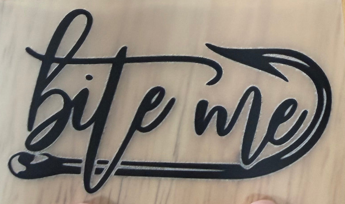 A hand points toward a small clear sheet with a black cursive 'bite me' graphic, with another sheet and a larger curved sheet visible above on a wooden tabletop.