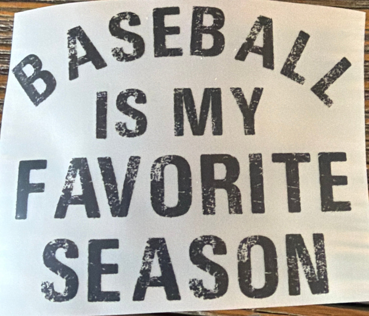 A white die-cut sticker with the text 'BASEBALL IS MY FAVORITE SEASON' in distressed black lettering, shown on a brown wood-grain surface.