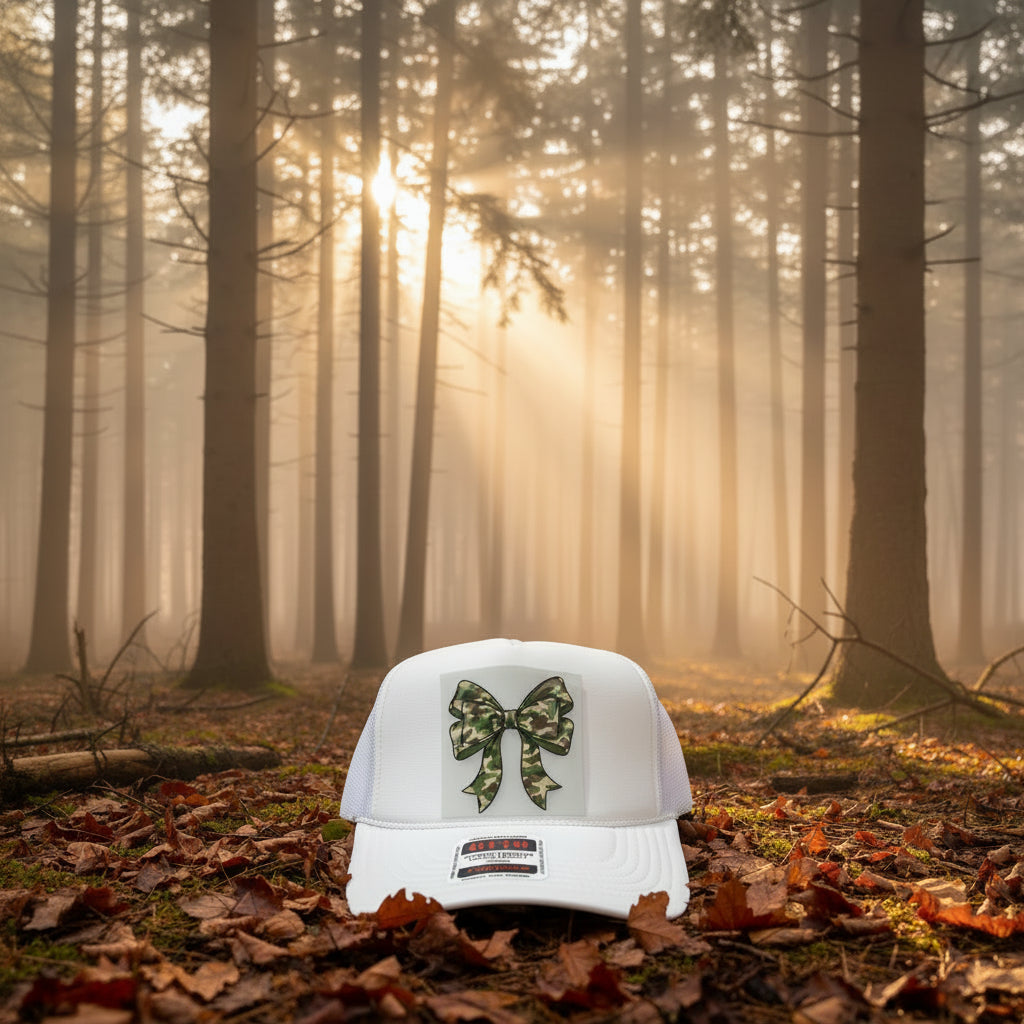 A white trucker hat with a green camouflage bow graphic on the front, resting on a wooden surface. The brim features a visible 'OTTO' brand sticker.