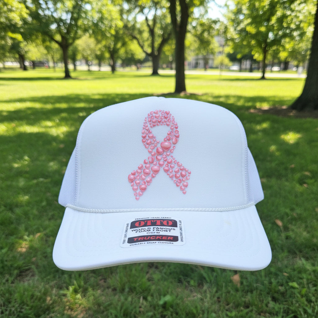 A white foam trucker hat with a pink awareness ribbon on the front, made of patterned pink foam. The hat has white mesh side/back panels and a visible 'OTTO' brand sticker on the brim.
