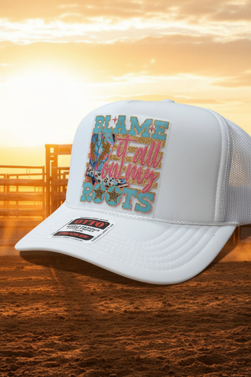 A white trucker hat with a colorful front patch reading 'BLAME it all on my ROOTS' and a visible Ottio foam fabric sticker on the brim, photographed on a wooden surface.