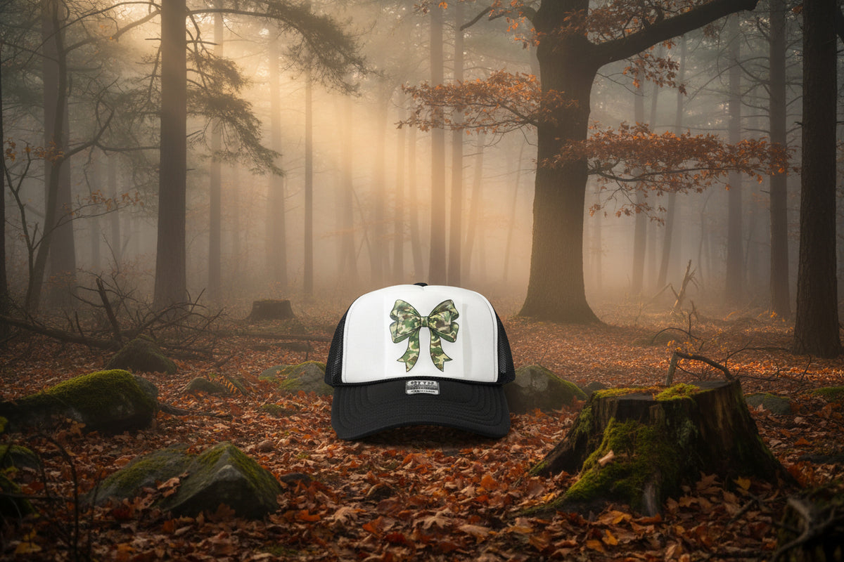 A white and black trucker hat with a green camouflage bow graphic on the front panel, displayed on a wooden surface.