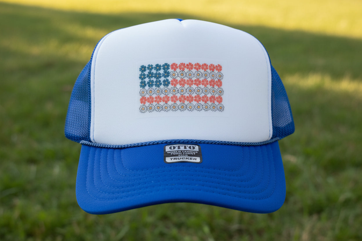 A blue and white trucker hat with a white front panel, blue mesh sides, and a blue brim. The front panel features a decorative American flag made of blue, red, and white daisy flowers.