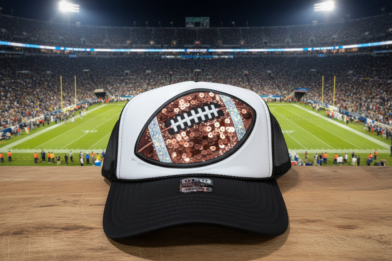 A black and white trucker hat with a large football graphic on the front panel. The football graphic features rose gold sequins, white laces, and light blue accents.