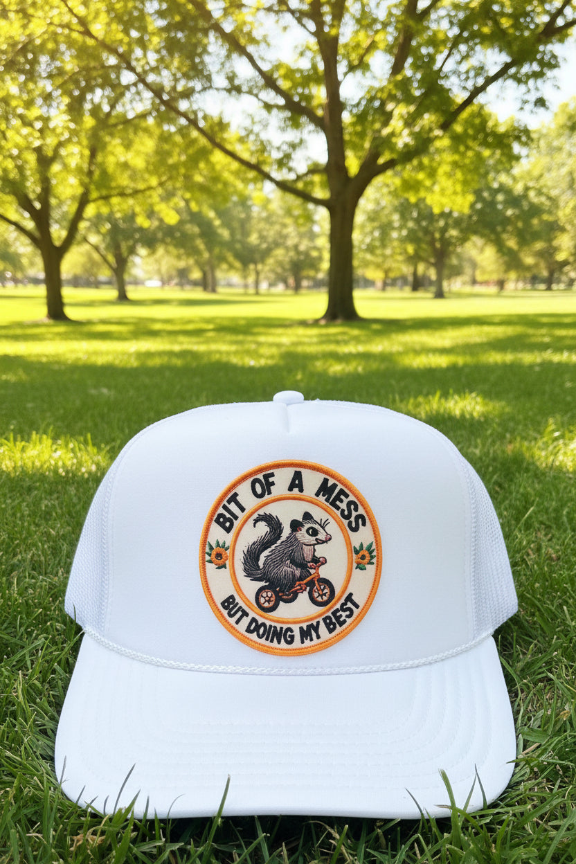 A white trucker hat with a circular embroidered patch on the front featuring a squirrel on a tricycle and the text 'BIT OF A MESS BUT DOING MY BEST.' A sewn-on OTTO brand sticker is visible on the brim.