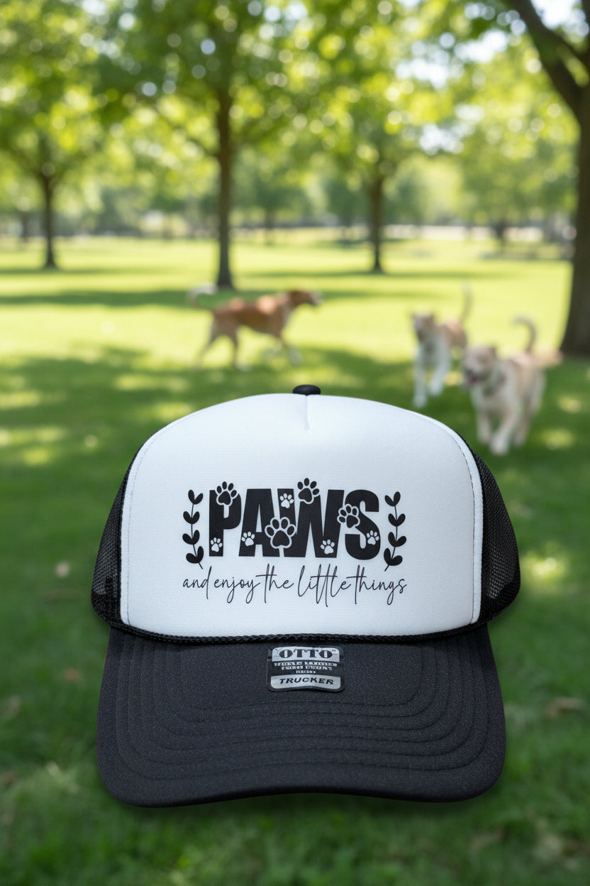 A black and white trucker hat with a white front panel and black mesh sides. The front panel features a graphic that reads 'PAWS and enjoy the little things' with paw prints and floral accents.