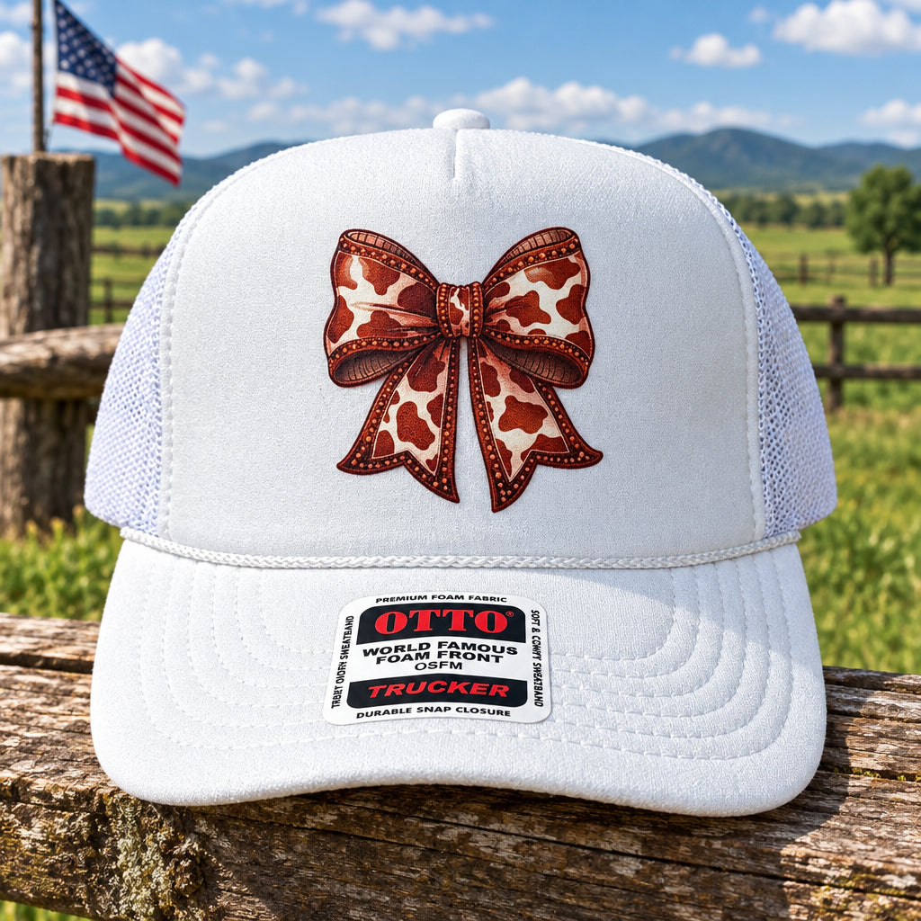 A white trucker hat with a mesh back, featuring a printed red and white cow-print bow graphic on the front. The brim has a visible 'OTTO' brand sticker.