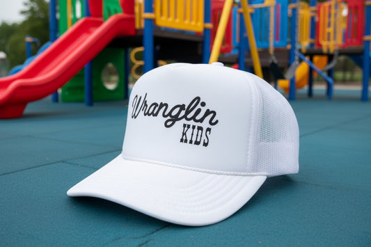 A white Wrangler Kids trucker hat with black script lettering on the front, resting on a wooden table.
