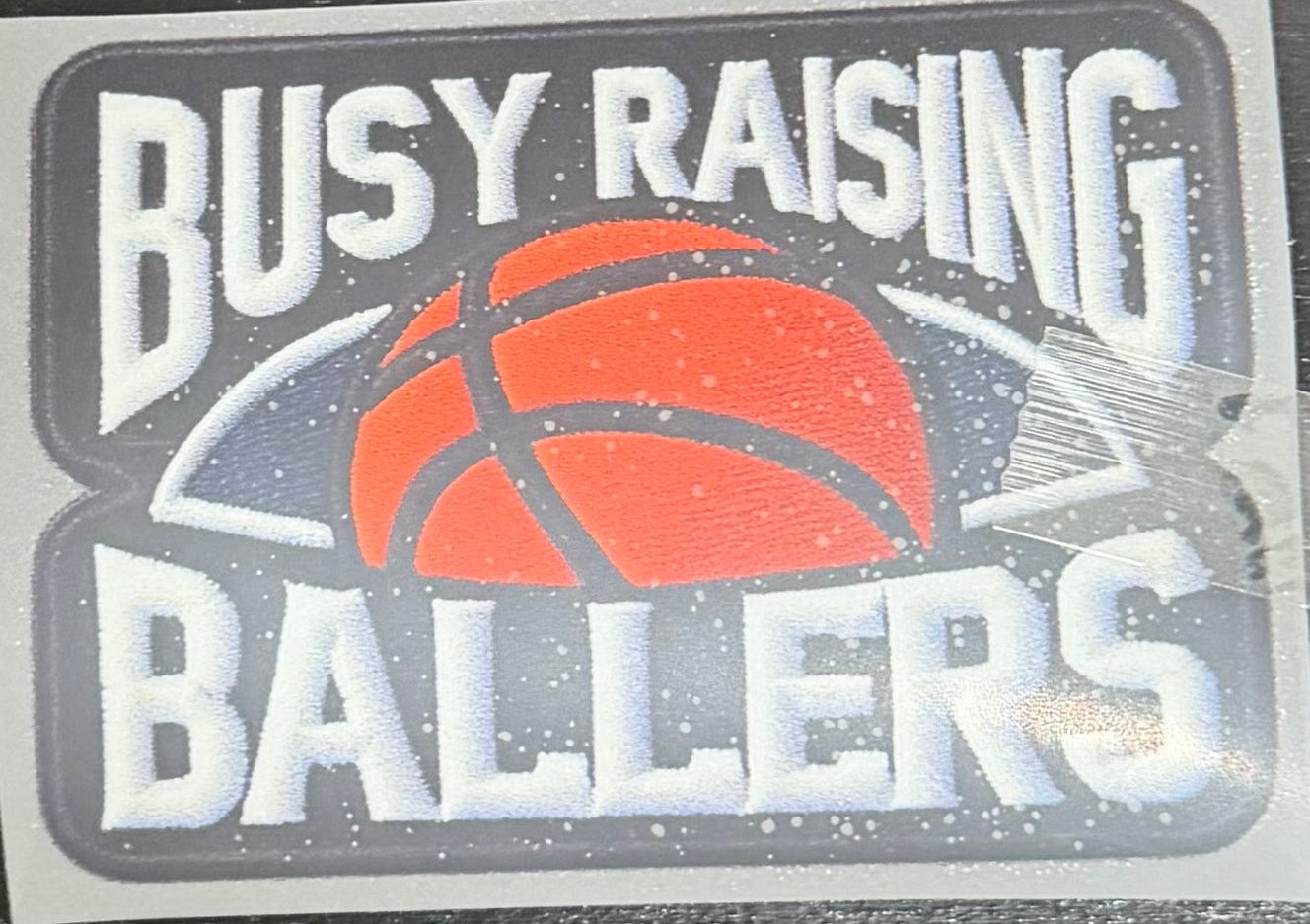 A DTF hat print with the text 'BUSY RAISING BALLERS' and a basketball graphic, placed on a dark wooden surface. 