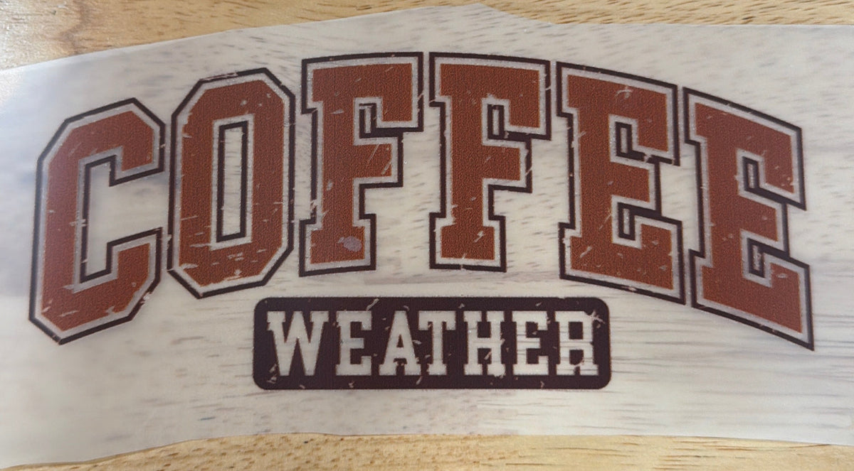 A vinyl decal sticker featuring the text 'COFFEE WEATHER' in an arched collegiate font, shown on a piece of clear tape over a light wood tabletop. A person's legs and shoes are visible at the bottom of the frame.