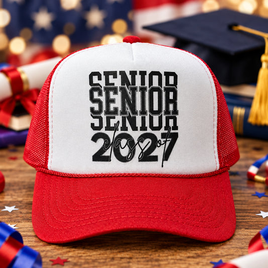 A red and white trucker hat with 'SENIOR SENIOR SENIOR 2027' printed in black on the front, displayed on a wooden table with two other hats in the background.