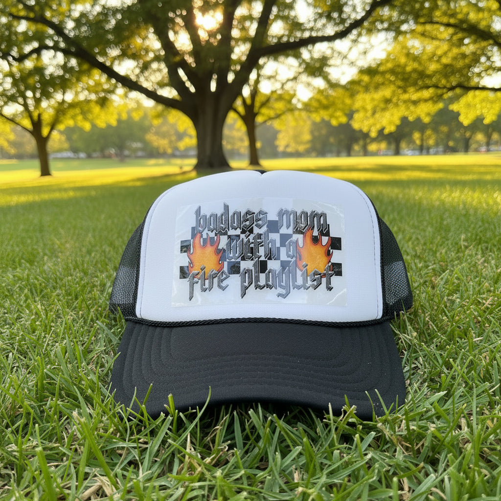 A black and white trucker hat with a custom graphic on the front panel that reads 'badass mom with a fire playlist' in gothic lettering with orange flames and a black-and-white checkered pattern. The hat has a black brim and black mesh back, with an Otto Trucker brand sticker on the brim.