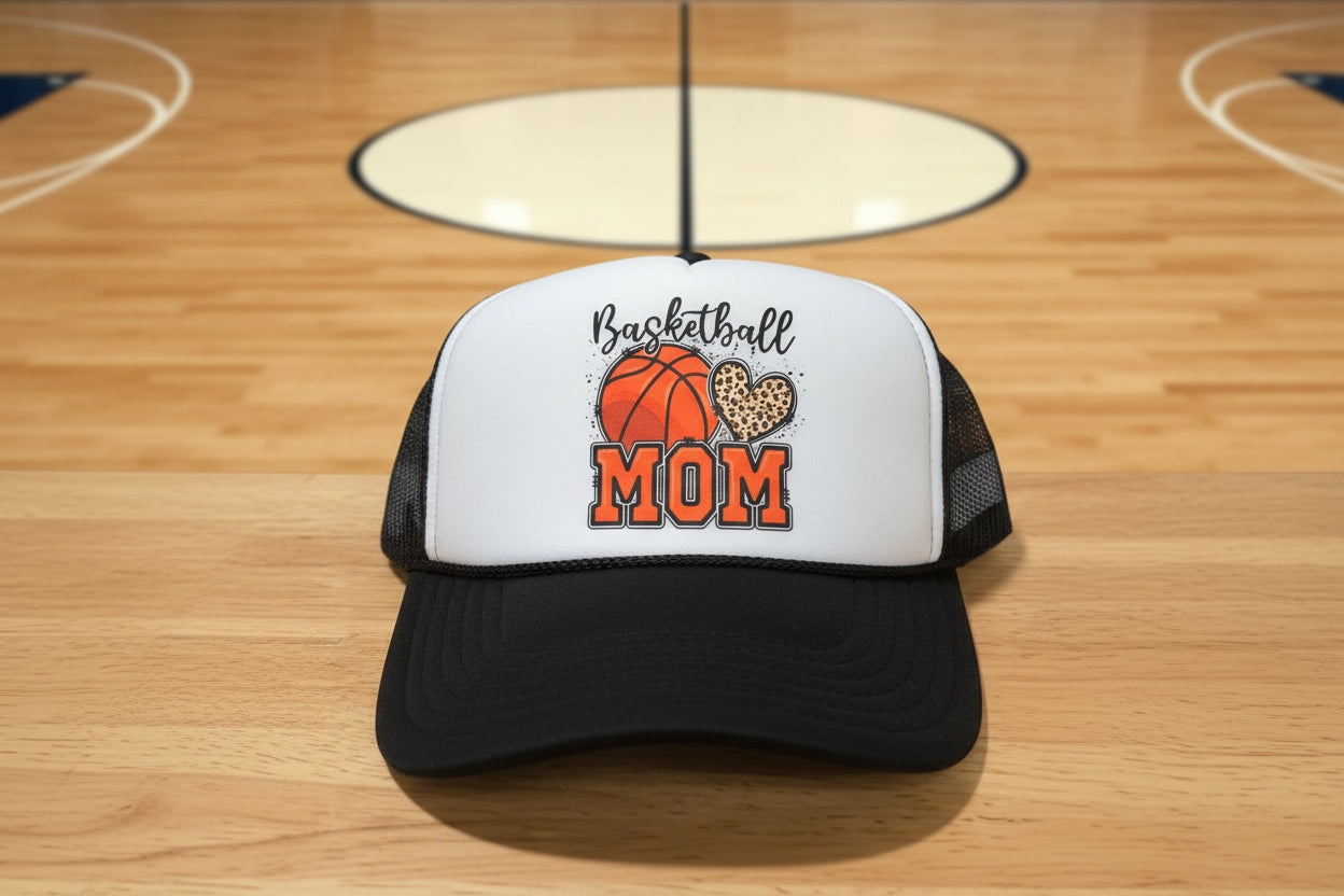 A black and white trucker hat with a custom 'Basketball MOM' graphic on the front panel, resting on a wooden table.