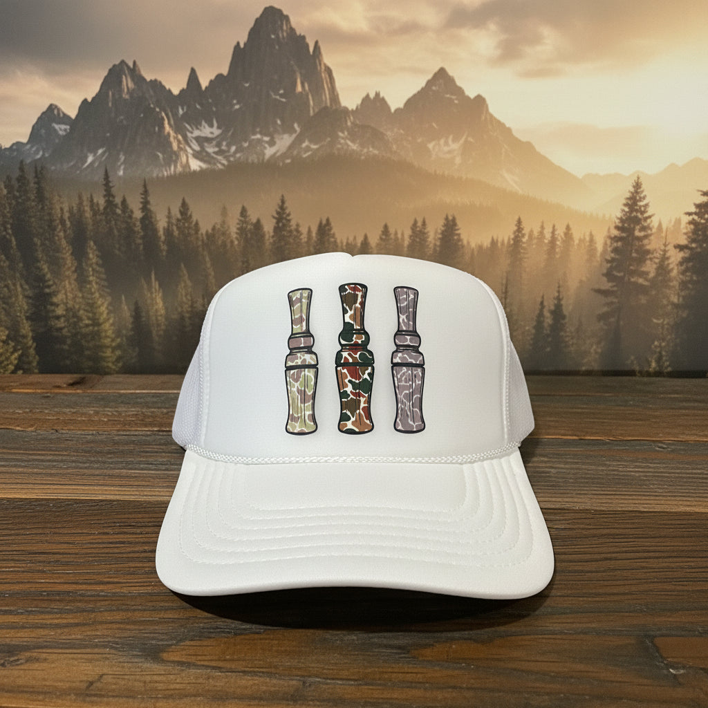 A white trucker hat with mesh side panels, featuring three camouflage duck-call graphics on the front and an OTTO Trucker label on the brim, displayed on a wooden surface.