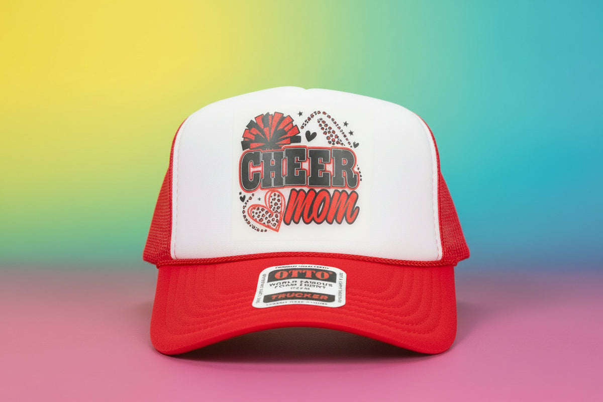 A red and white trucker hat with a 'CHEER Mom' graphic on the front, displayed on a black table against a wooden wall background.