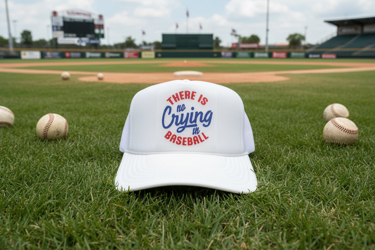 A white trucker hat with a front patch reading 'THERE IS no Crying in BASEBALL' in red and blue lettering, photographed on a dark wooden surface.