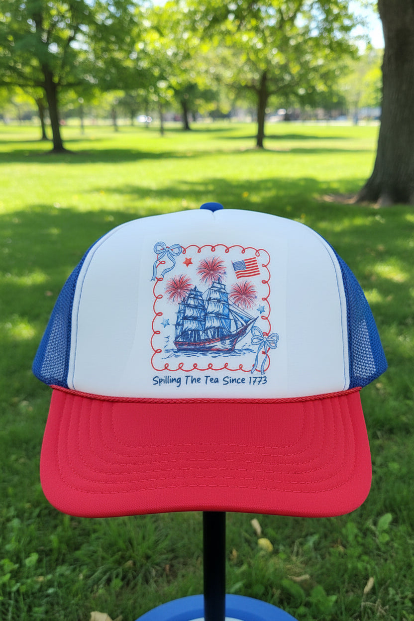 A red and blue trucker hat with a white front panel featuring a patriotic nautical graphic of a sailing ship, fireworks, an American flag, and the text 'Spilling The Tea Since 1773'.