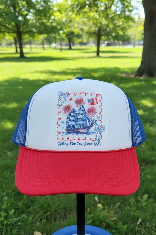 A red and blue trucker hat with a white front panel featuring a patriotic nautical graphic of a sailing ship, fireworks, an American flag, and the text 'Spilling The Tea Since 1773'.