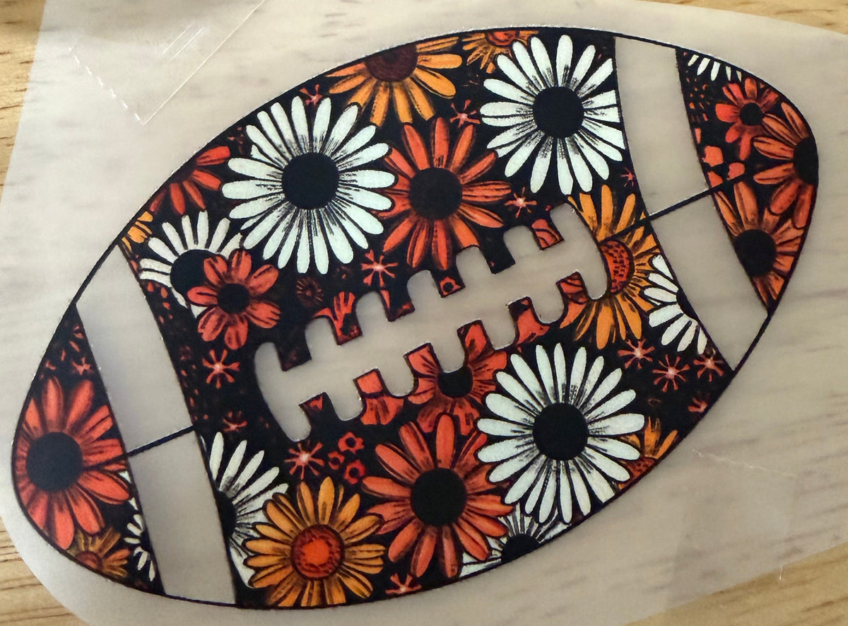 A football-shaped decal or sticker with a floral pattern in orange, white, and black, placed on a piece of clear tape over a light wood table.