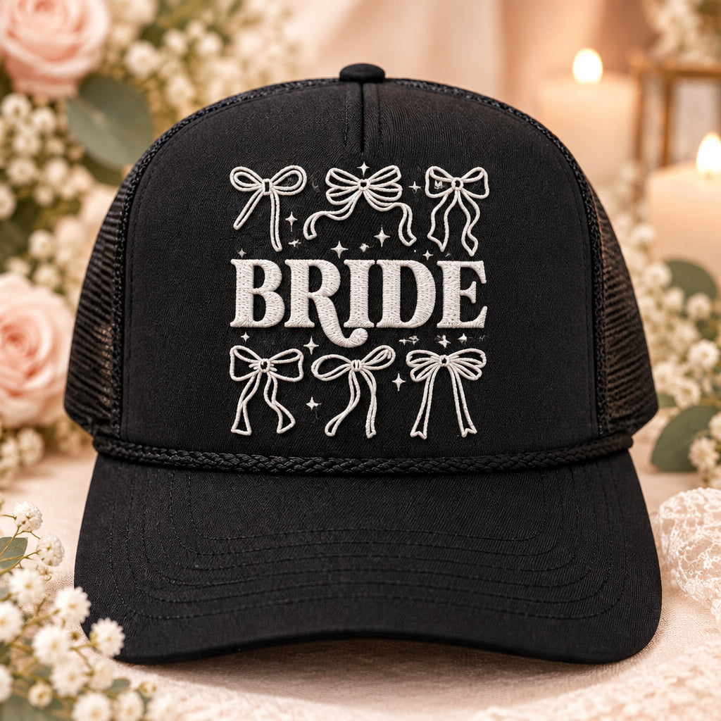 A black trucker hat with a gray graphic on the front that reads 'BRIDE' with bow illustrations, displayed on a wooden table. The hat has a visible Ottobaro sticker on the brim.