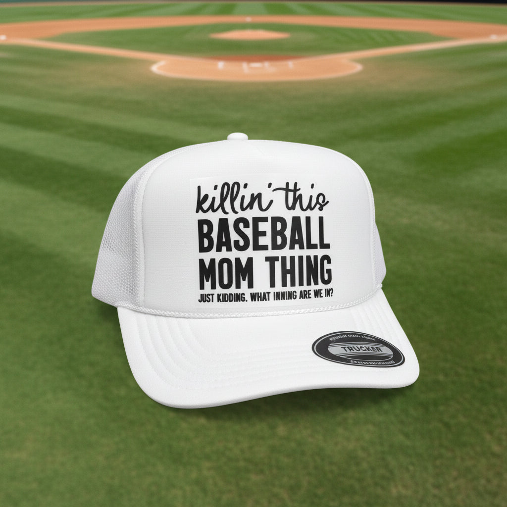 A white trucker hat with black text reading 'killin' this BASEBALL MOM THING' and smaller text below, photographed on a dark tabletop against a dark wood background. A visible patch on the brim reads OTTO World Famous Foam Front 1969 Trucker.