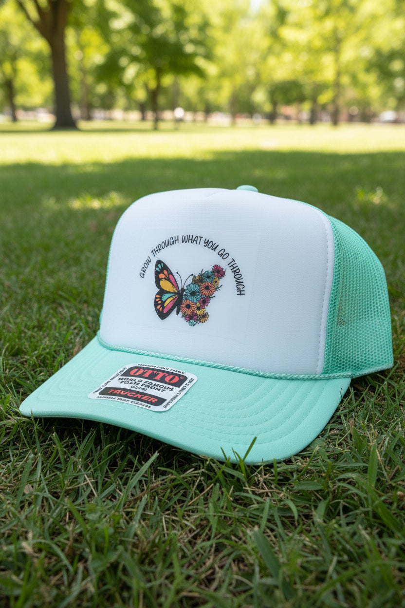 A mint green and white foam trucker hat with a butterfly graphic on the front panel. The hat features a white front panel with a colorful butterfly design and the text 'CROWN THROUGH WHAT YOU GO THROUGH' arched above it.