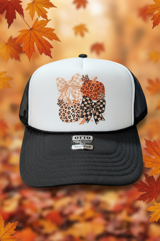 A black and white trucker hat with a custom fall-themed graphic on the front panel, featuring two patterned pumpkins and two checkered bows. The hat is displayed on a wooden surface.