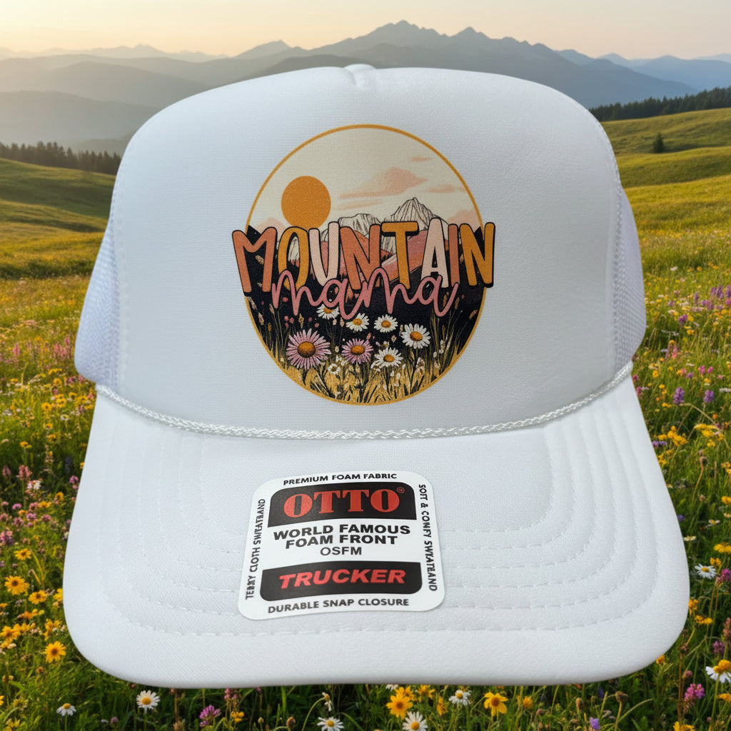 A white trucker hat with a circular 'Mountain Magic' graphic on the front, resting on a wooden table. The hat features a white structured front panel, white mesh side/back panels, and a white sticker on the brim that reads 'OLD WORLD FAMOUS YOUR FRONT LINE TRUCKER'.