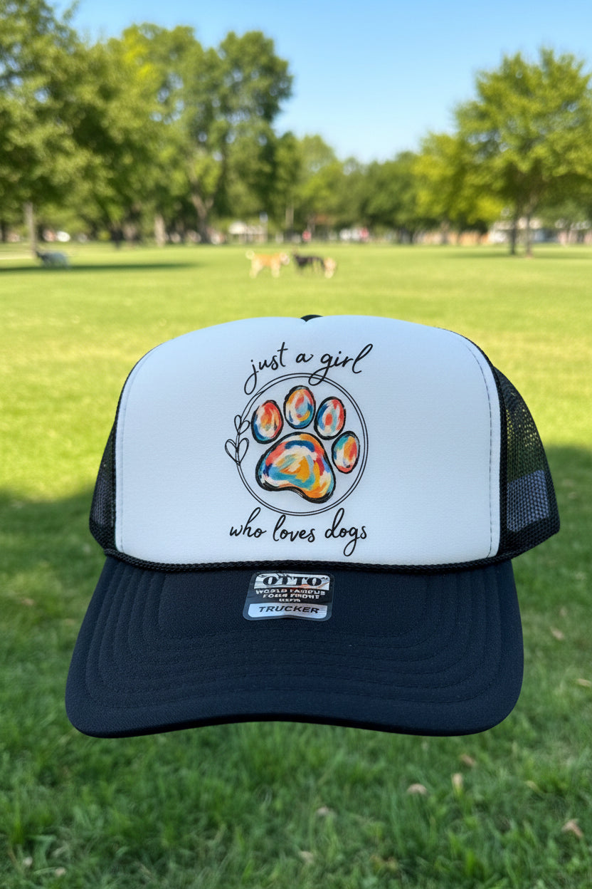 A black and white trucker hat with a colorful dog paw print graphic and the text 'just a girl who loves dogs' on the front panel.