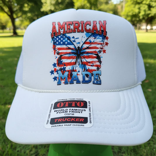 A white trucker hat with a patriotic American flag graphic on the front, resting on a wooden table. The hat features a red, white, and blue butterfly design with the words 'AMERICAN' and 'Patriot'.