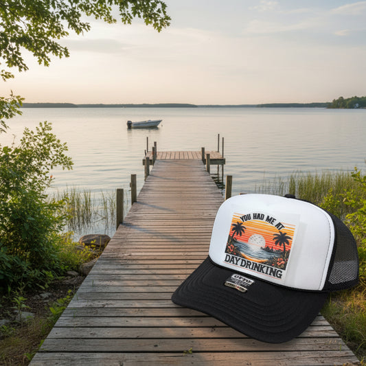 A black and white trucker hat with a retro sunset graphic on the front panel that reads 'YOU HAD ME AT DAY DRINKING'. The hat has a black brim, black mesh sides, and a white front panel.
