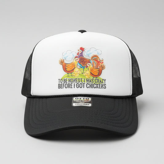 A white and dark gray trucker hat with a graphic of colorful chickens and the text 'TO BE honest I WAS CRAZY BEFORE I GOT CHICKENS' displayed on a black table against a wooden background.
