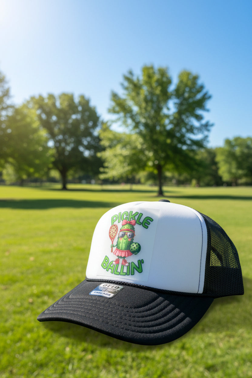 A black and white trucker hat featuring a front patch with a cartoon cactus character holding a pickleball racket and the text 'PICKLE BALLIN''. A metal badge on the brim reads 'OMG WORLD FAMOUS PICKLE BALLIN' TRUCKER'.