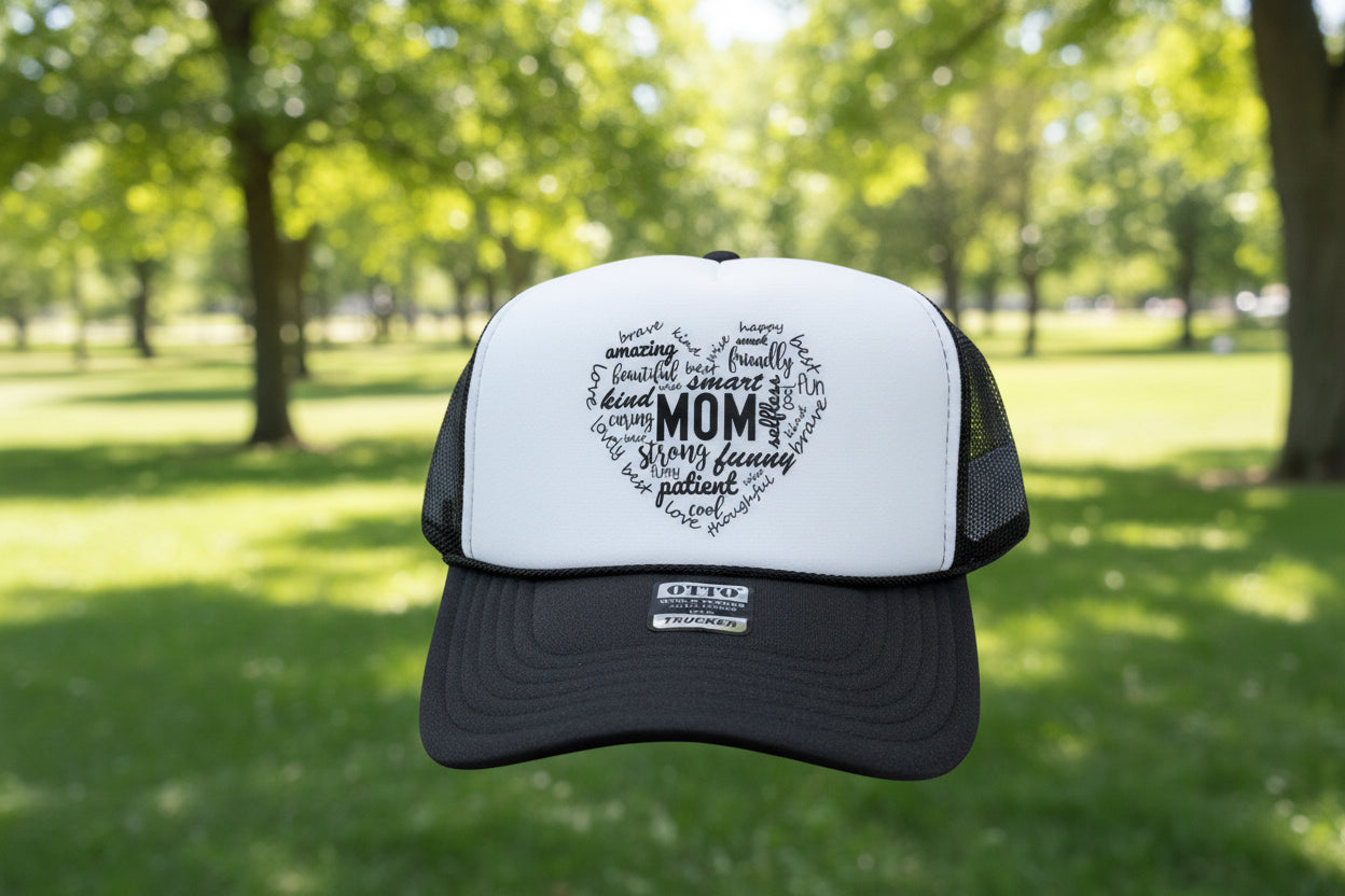 A black and white trucker hat with a heart-shaped graphic on the front panel that reads 'MOM' surrounded by positive adjectives. The hat has a black brim, black mesh back, and a visible OTTO brand sticker on the front.