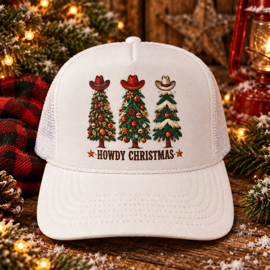A white trucker hat with a 'Howdy Christmas' graphic on the front panel, featuring three decorated Christmas trees with cowboy hats. The hat has a visible Otto brand sticker on the brim and is photographed on a wooden surface.