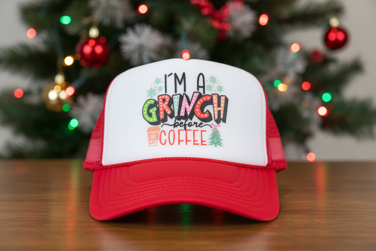 A red and white trucker hat with a festive graphic that reads 'I'M A GRINCH before COFFEE' on the front panel, resting on a wooden surface.