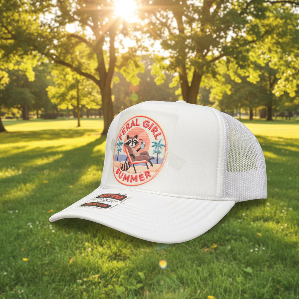 A white trucker hat with a circular 'Feral Girl Summer' raccoon patch on the front and an OTK Trucker sticker on the brim, set against a dark wood background.