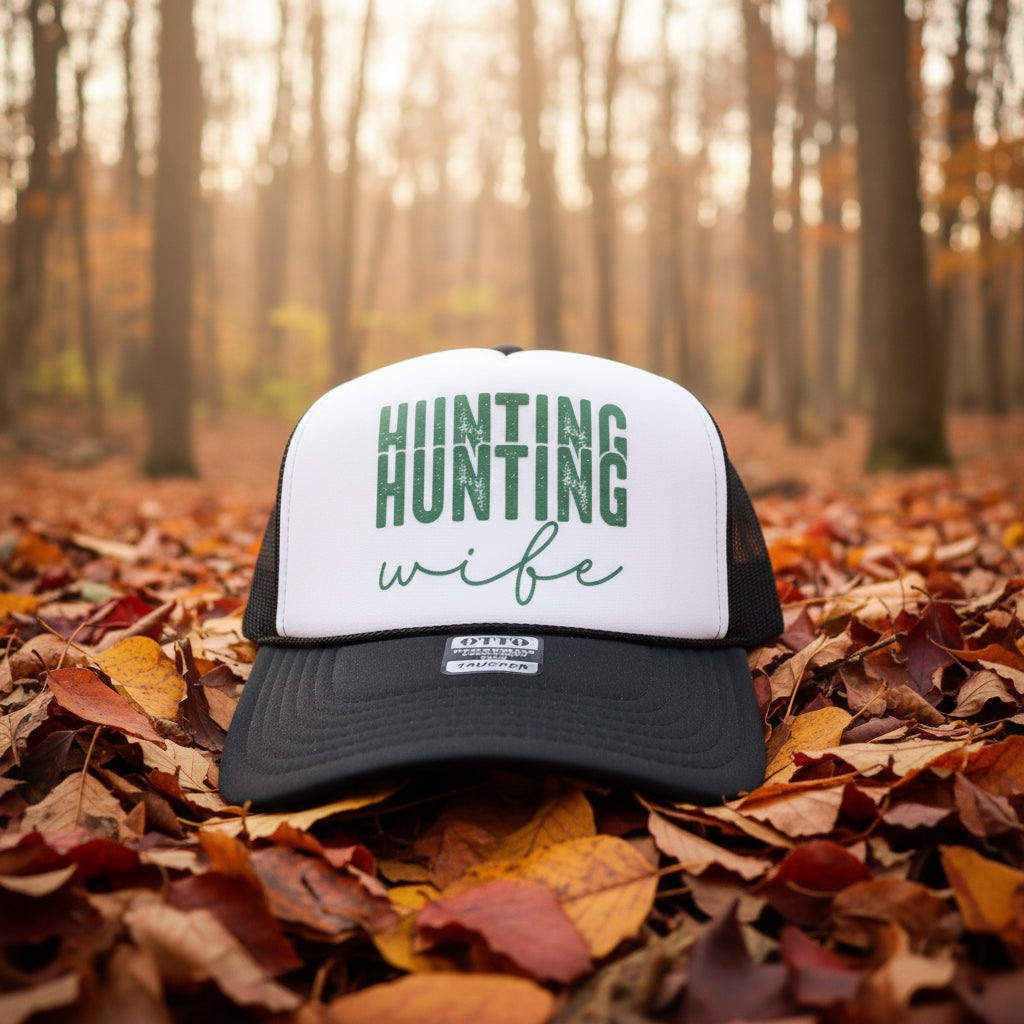 A black and white trucker hat with 'HUNTING wife' printed on the front, resting on a rustic wooden surface.