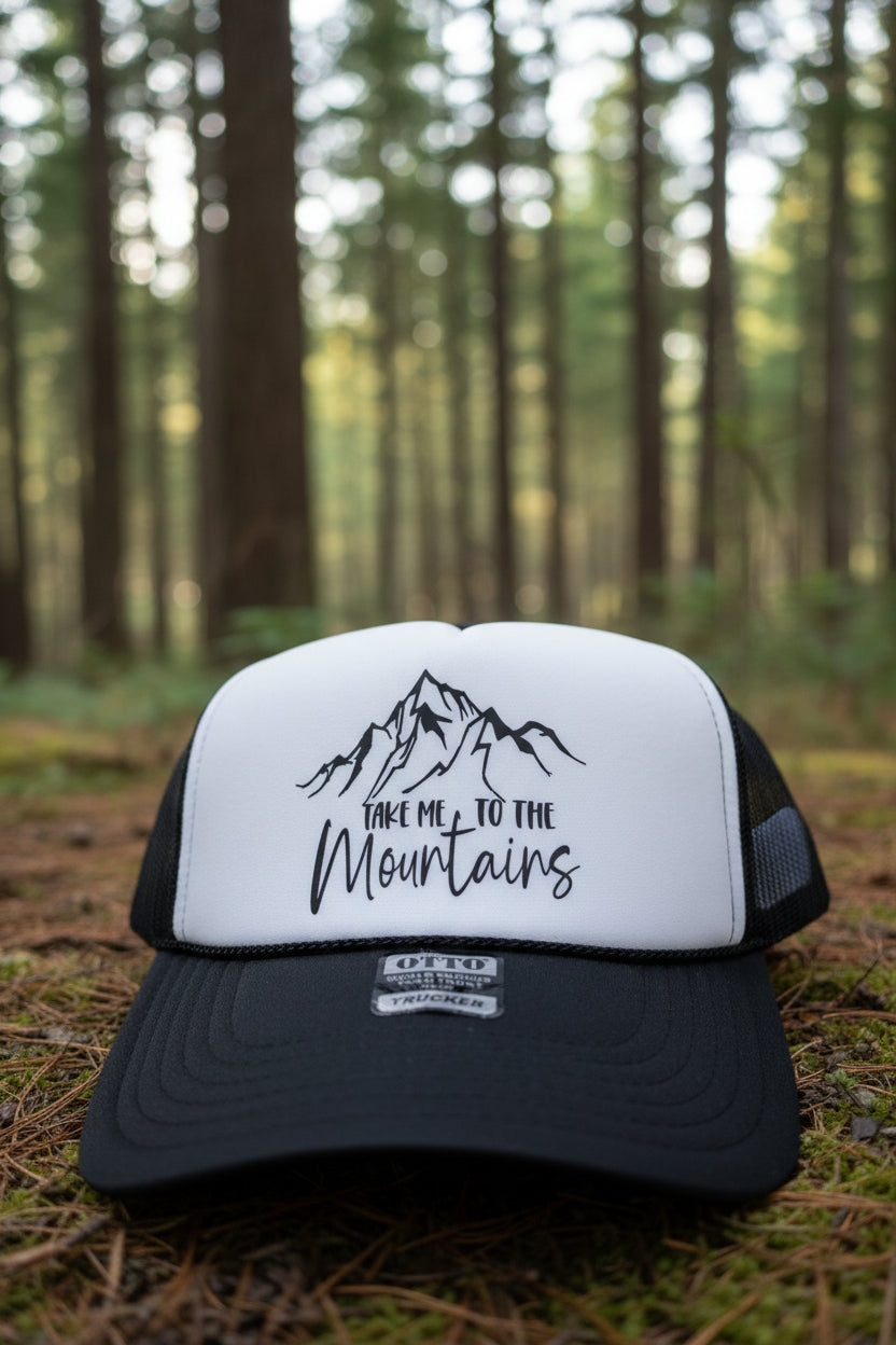 A black and white trucker hat with a mountain graphic and the text 'TAKE ME TO THE Mountains' on the front panel. The brim is black, and the mesh back is black.