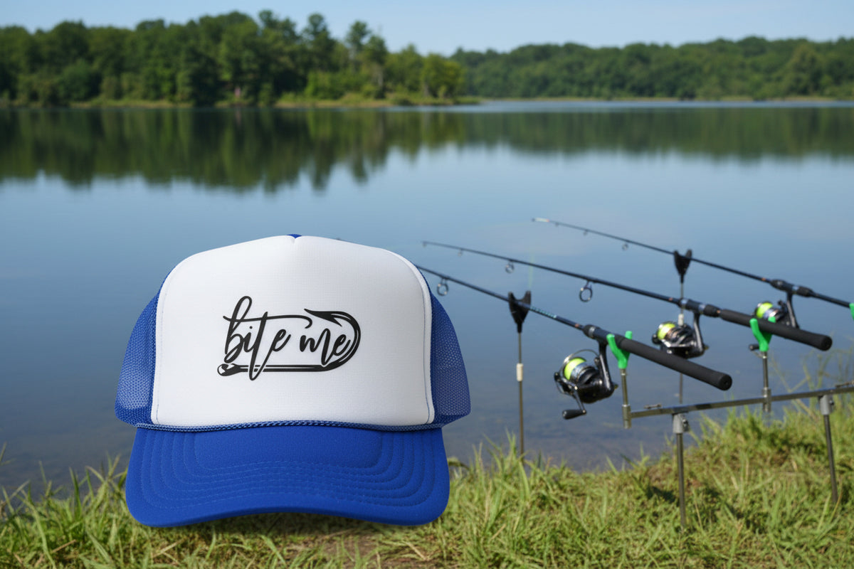 A blue and white trucker hat with a white front panel, royal blue brim, and mesh back. The front panel features a black graphic that reads 'bite me' in script.