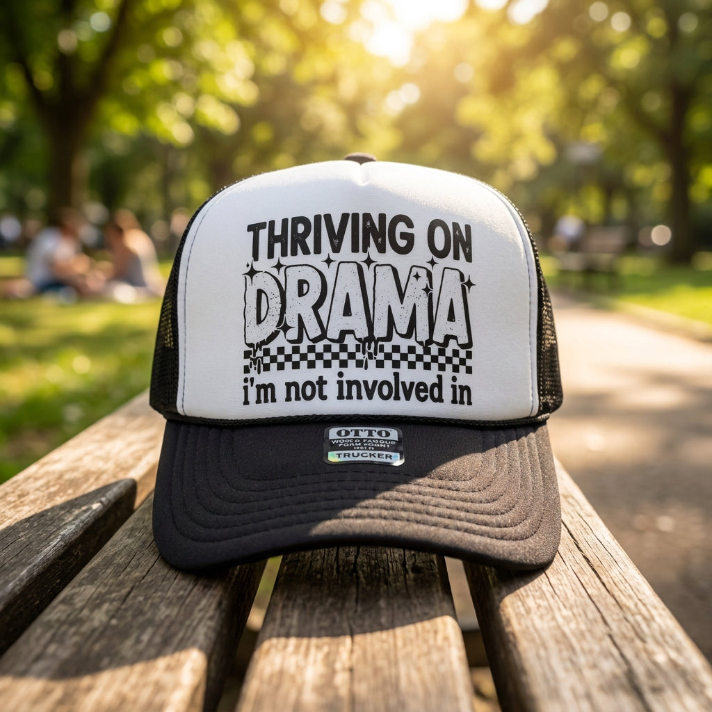 A black and white trucker hat with the text 'THRIVING ON DRAMA i'm not involved in' printed on the front panel, resting on a wooden surface.