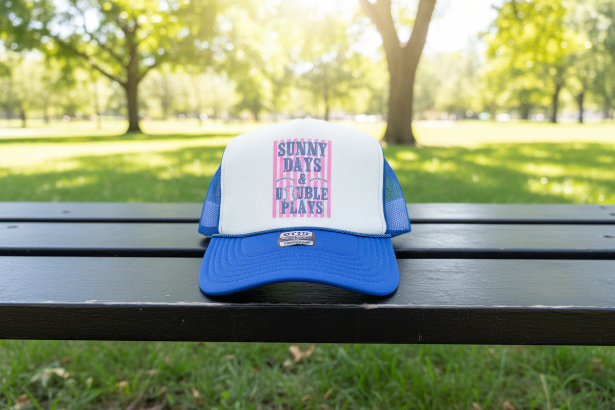 A blue and white trucker hat with a pink and white striped front patch that reads 'SUNNY DAYS & DOUBLEPLAYS'. The hat is resting on a dark wooden table against a dark wood-paneled wall.