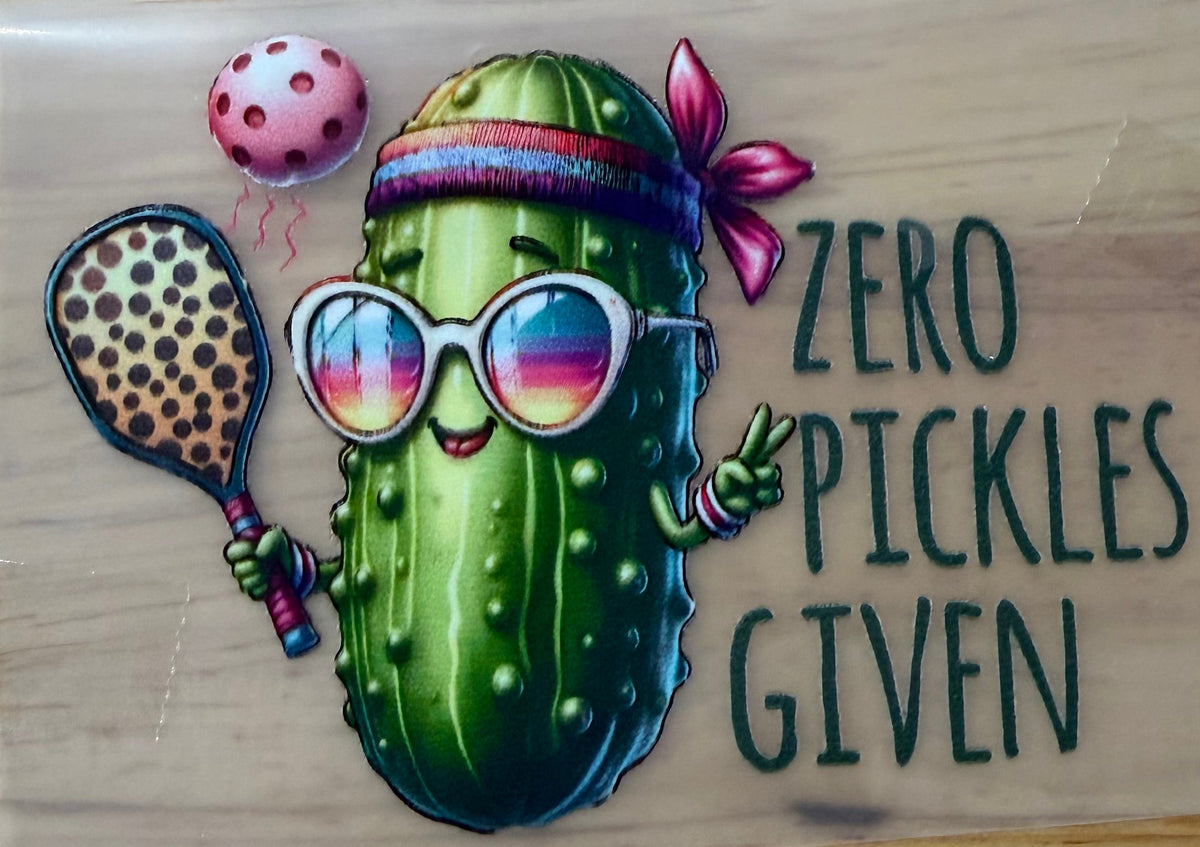 A rectangular printed sticker or decal on a wooden surface, featuring a cartoon pickle wearing sunglasses, a headband, and a pink bow, holding a racquet, with the text 'ZERO PICKLES GIVEN'.