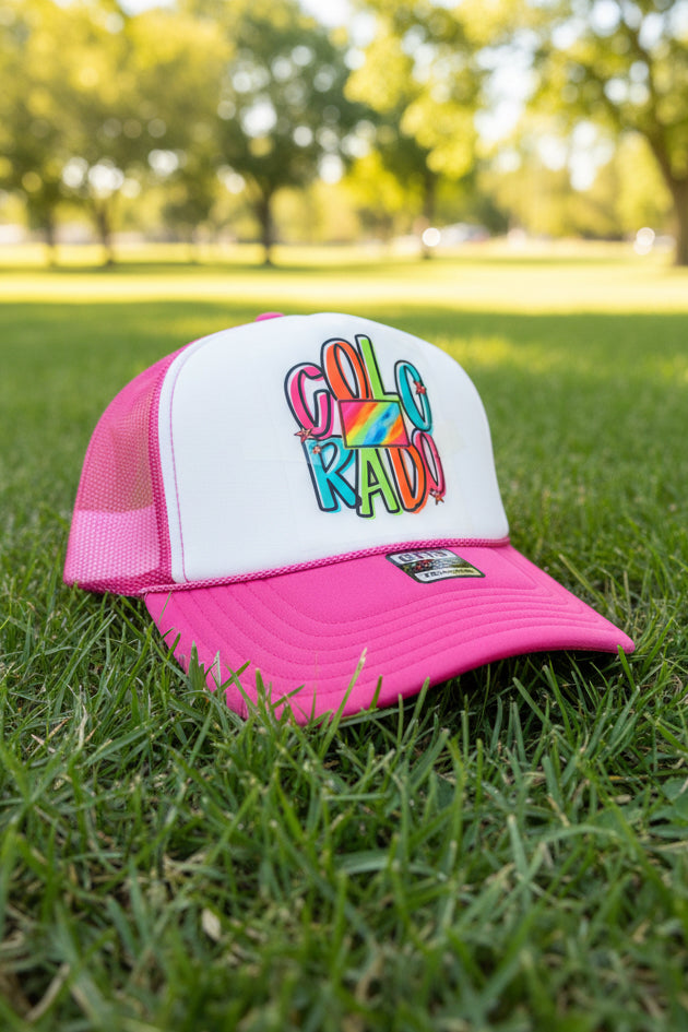 A pink and white trucker hat resting on green grass in an outdoor park setting. The front of the cap features a white panel with a colorful graphic that reads 'Colorado' in rainbow letters over a heart, and the mesh back and brim are bright pink.