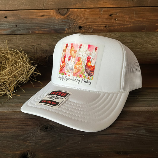 A white trucker hat with a colorful chicken-themed graphic on the front and an OTTO Trucker sticker on the visor, set against a dark wood background.