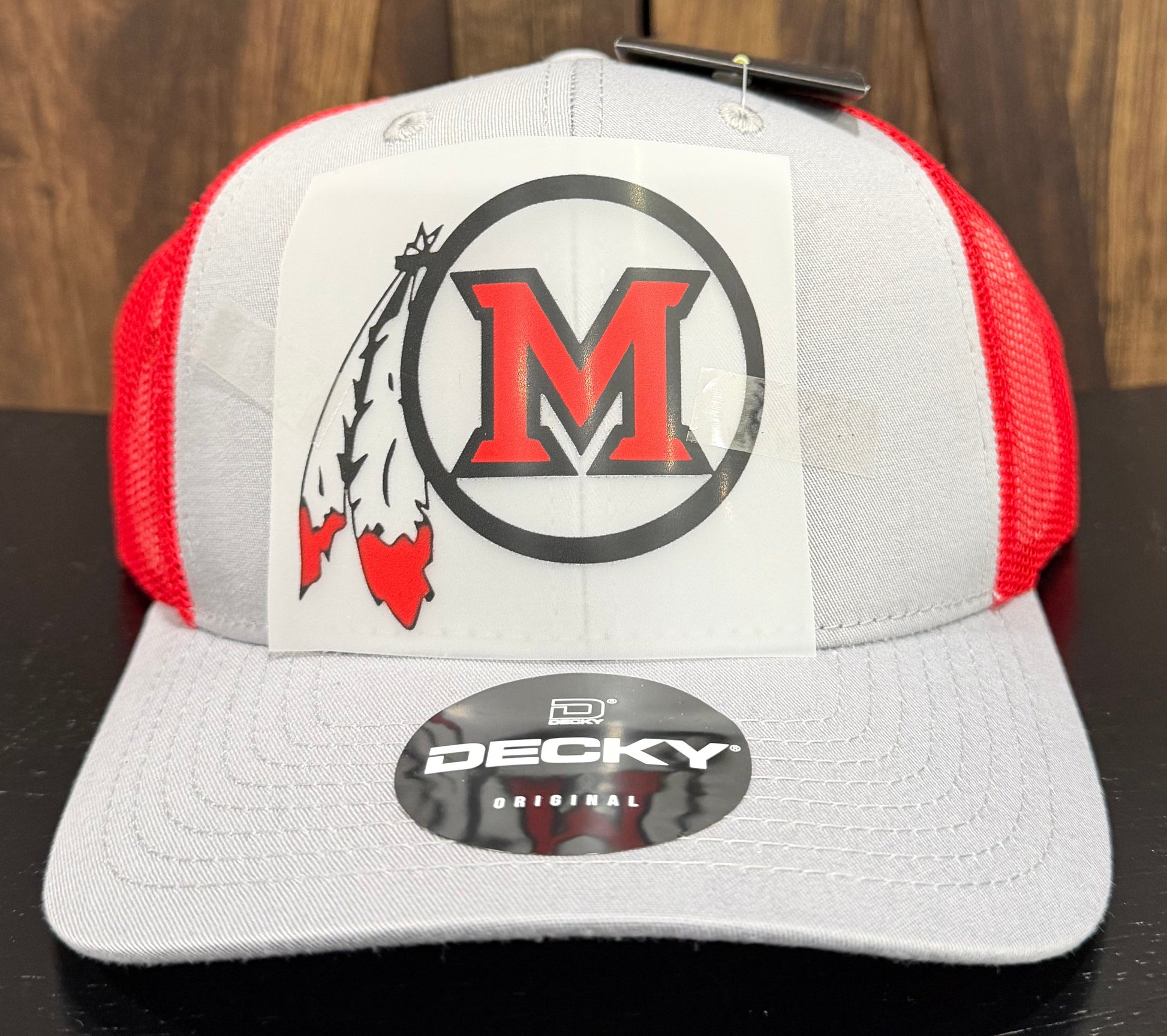 A gray and red trucker hat with a white front patch featuring a red 'M' logo and a black-and-white feather graphic.