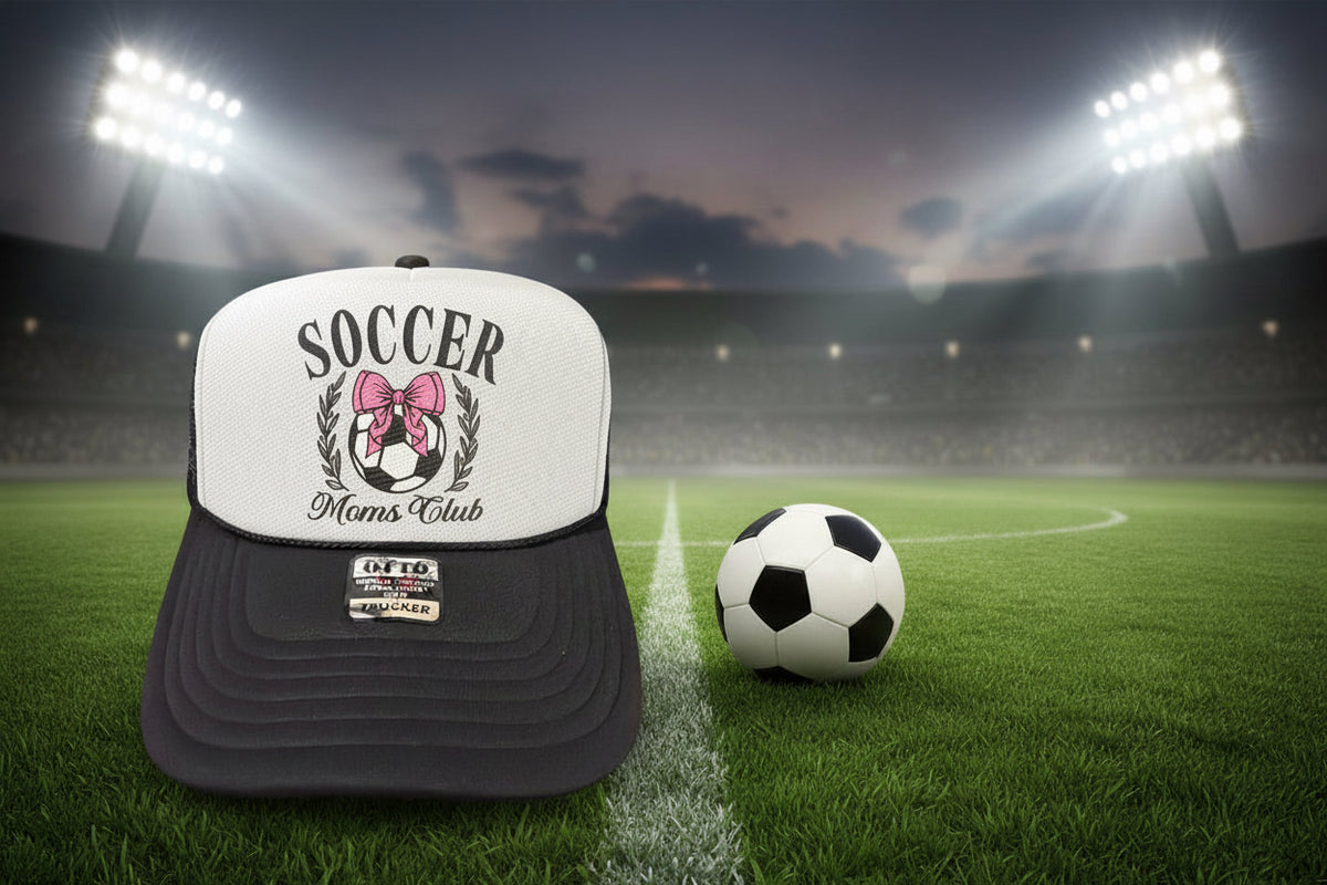 A black and white trucker hat with a custom 'Soccer Moms Club' graphic on the front panel, featuring a pink bow and soccer ball design. The hat is displayed on a rustic wooden surface.