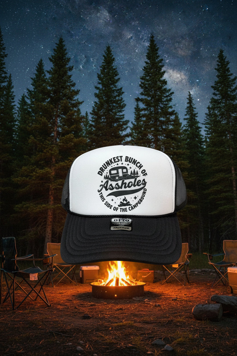 A black and white trucker hat with a white front panel and black mesh back, featuring a black graphic that reads 'DRUNKEST BUNCH OF Assholes THIS SIDE OF THE CAMPGROUND' around a camper illustration. A black brim has a visible 'OTTO' brand sticker.