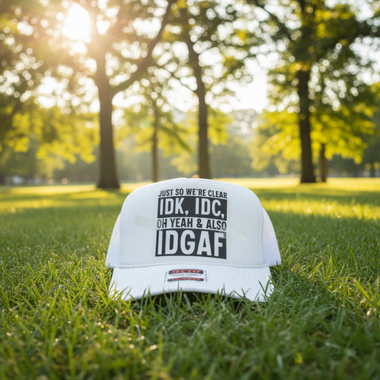 A white trucker hat with a printed graphic on the front that reads 'JUST SO WE'RE CLEAR IDK, IDC, OH YEAH & ALSO IDGAF' in black and white text. 