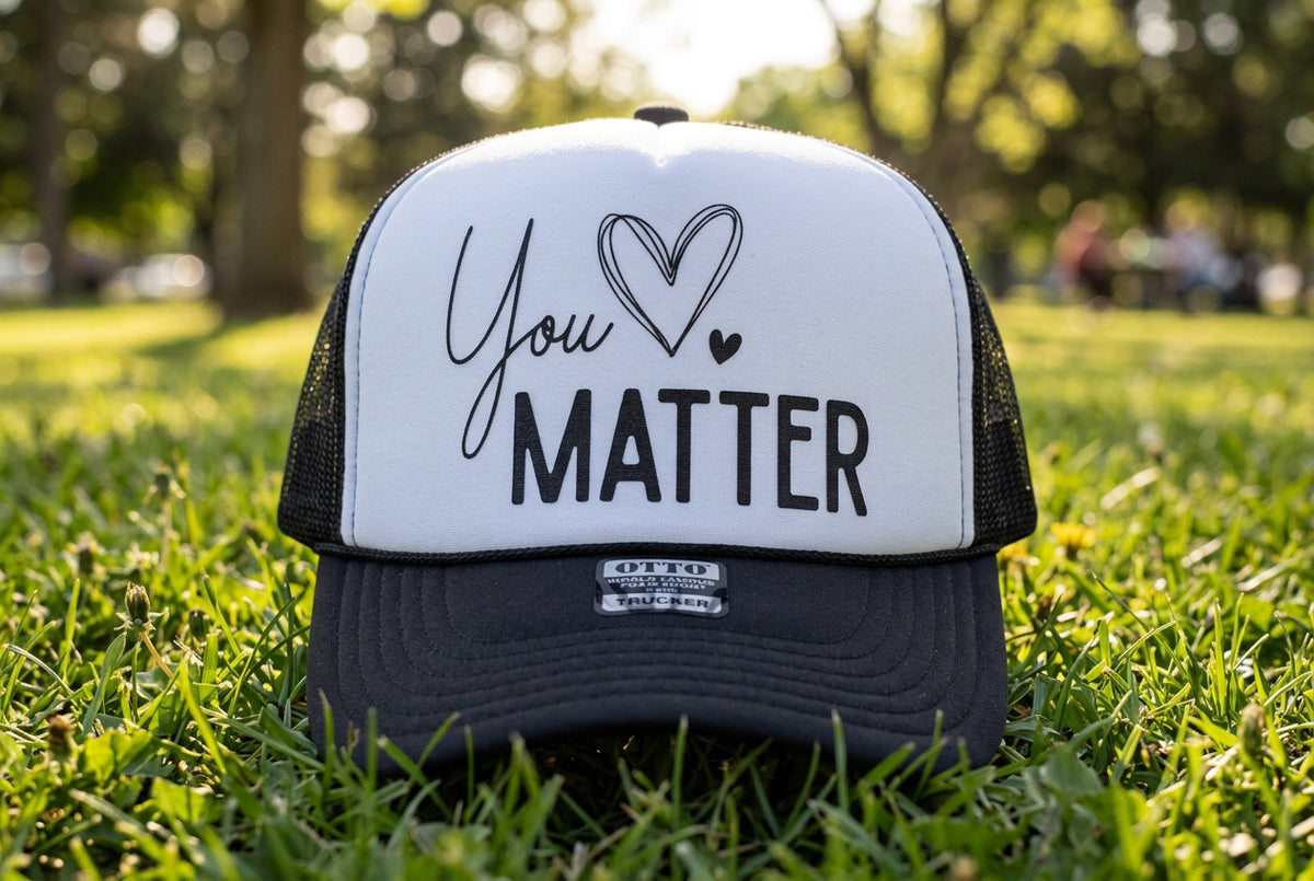 A black and white trucker hat with the words 'You' and 'MATTER' and a heart design printed on the front panel, resting on a wooden surface.